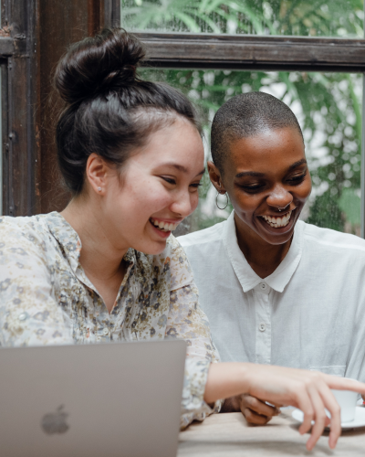 Image of asian woman and black woman sitting next to each other in front of a laptop and smiling at a coffee shop