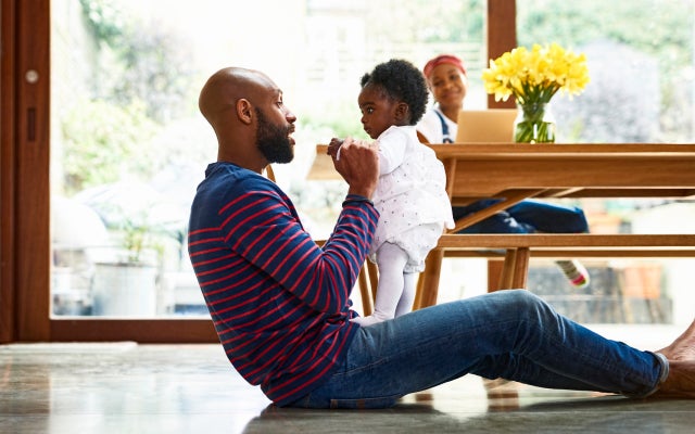Photo of black man sitting on the ground ground holding up daughter with wife on laptop at a table in the background