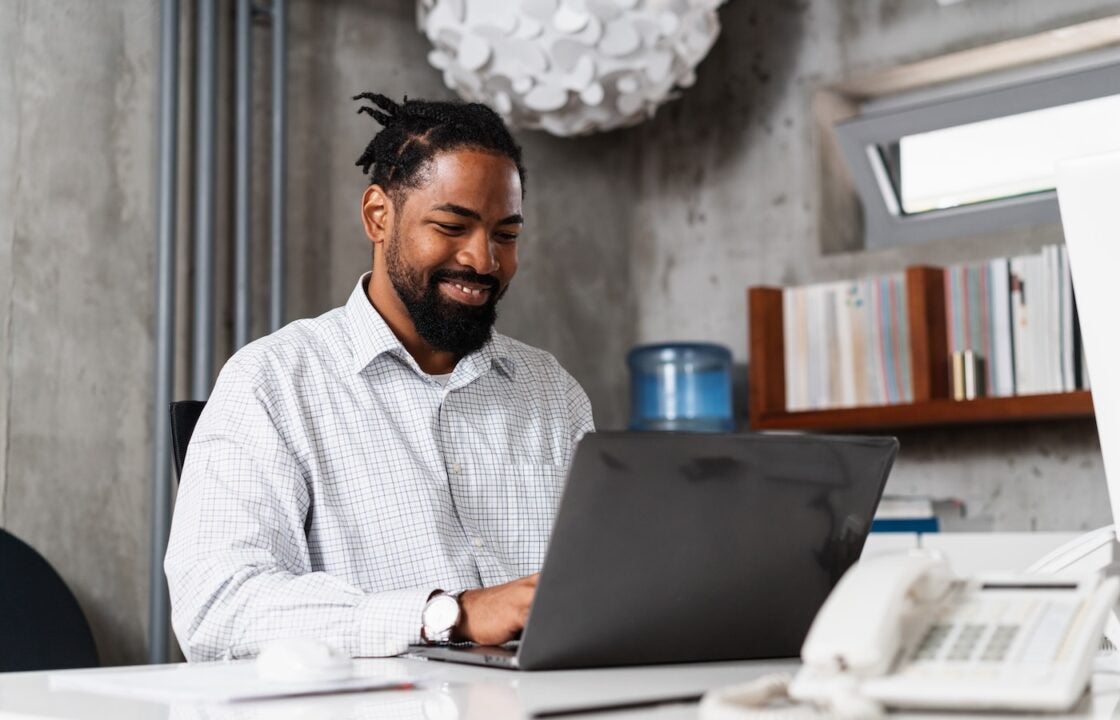 Smiling businessman sitting in an office, working through asset‑based employment checks on a laptop.