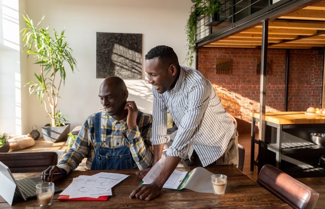 Two men working from home office together. One is sitting at the table in front of a laptop with documents and the other is standing next to him.