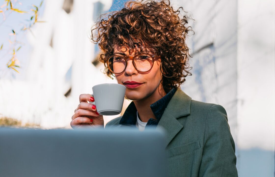 A businesswoman drinks coffee while working on a laptop outdoors.